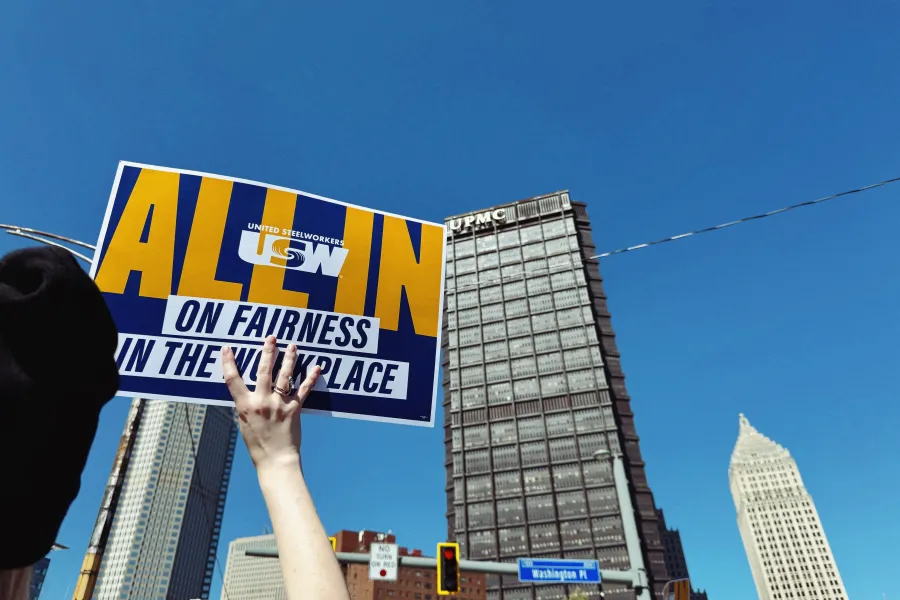 Arm holding up a flyer that says "All In on Fairness in the Workplace" with the buildings in the background of blue skies